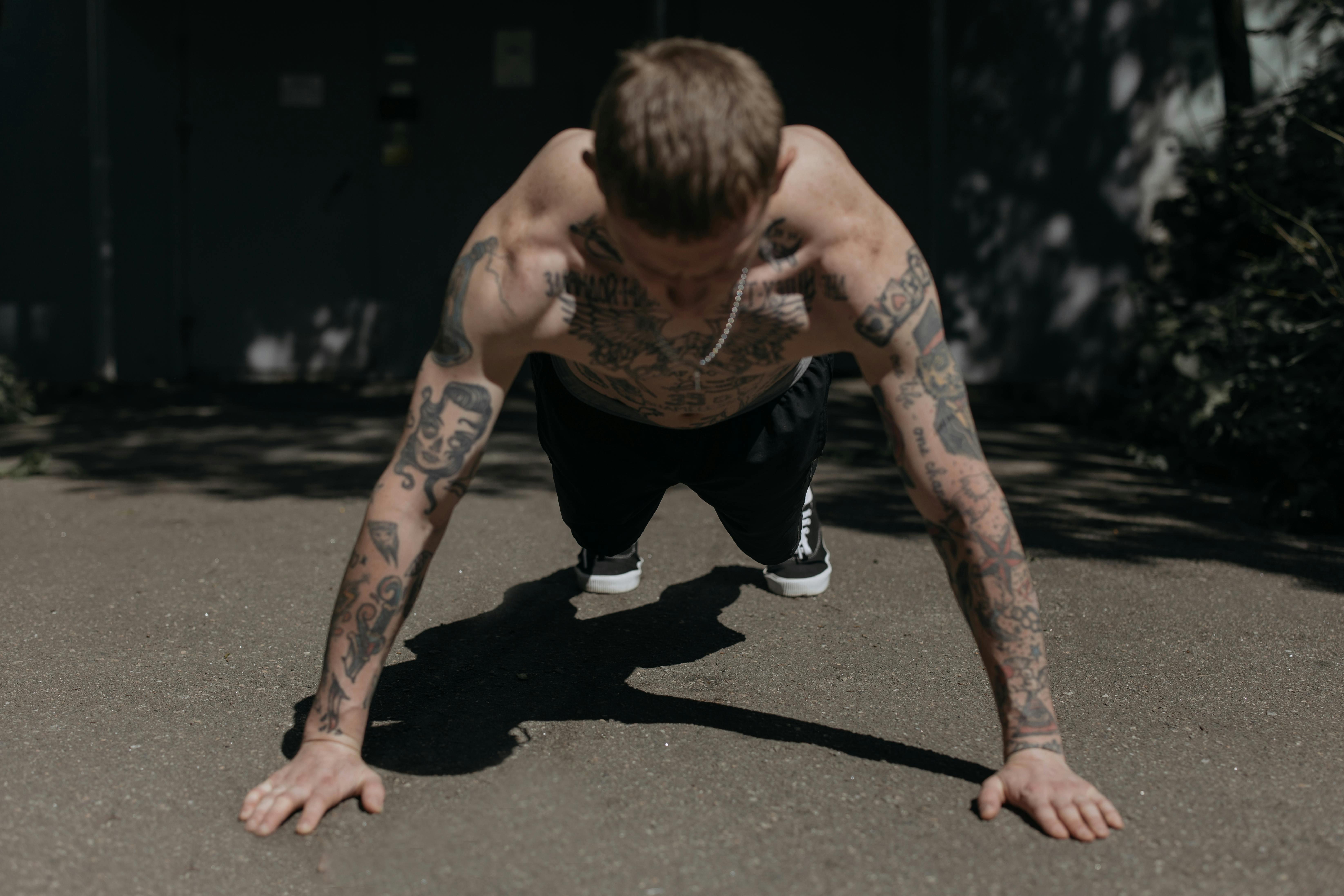 Shirtless tattooed man performing push-ups on a concrete path outdoors, showcasing fitness and endurance.