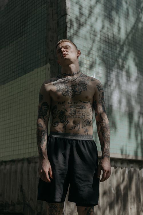Free Shirtless tattooed man standing outdoors in sunlight, showcasing artistic body art against a concrete backdrop. Stock Photo