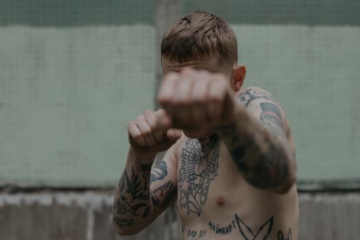 A tattooed male boxer training outdoors, throwing a punch against a concrete wall backdrop.