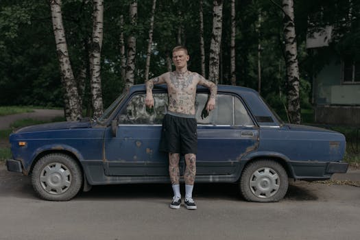 A tattooed man in shorts leans against a vintage car outdoors surrounded by trees.