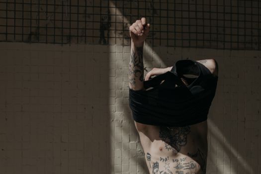 A tattooed man partially dressed in a black tank top, captured in moody indoor lighting.