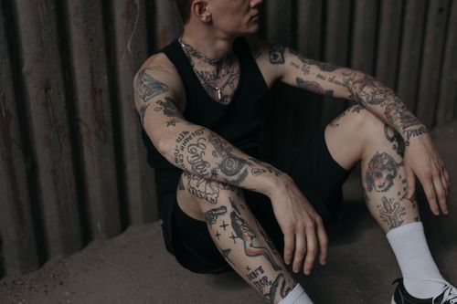 Free Close-up of a tattooed man sitting against a metal wall, wearing dark attire. Stock Photo