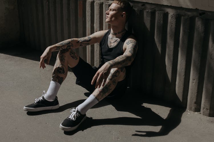 A Tattooed Man In Black Tank Top Sitting On The Ground While Leaning On The Wall