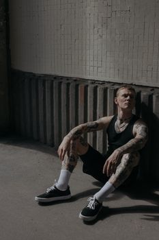 一名纹身男子身穿黑色背心，坐在水泥地板上，看起来若有所思