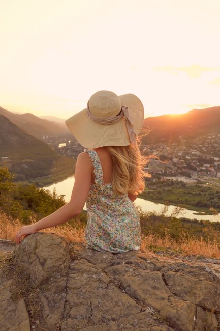 A woman in a floral dress and hat sits on a rock, enjoying a serene sunset view from a hillside.