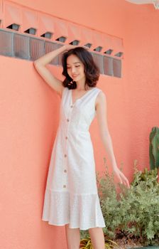 一位身穿白色连衣裙的时尚年轻女子，倚在粉红色的墙上，脸上带着安详的微笑