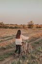 Back view of faceless lady with dark hair rolling bicycle on grassy road in countryside in summer evening in nature