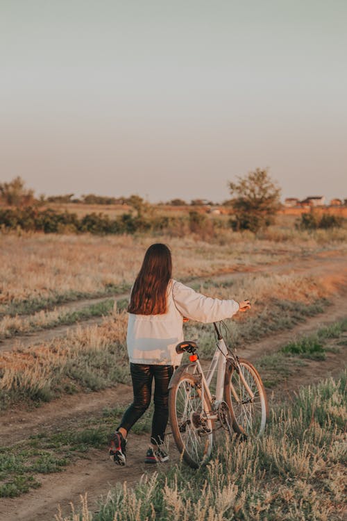 Free Back view of faceless lady with dark hair rolling bicycle on grassy road in countryside in summer evening in nature Stock Photo