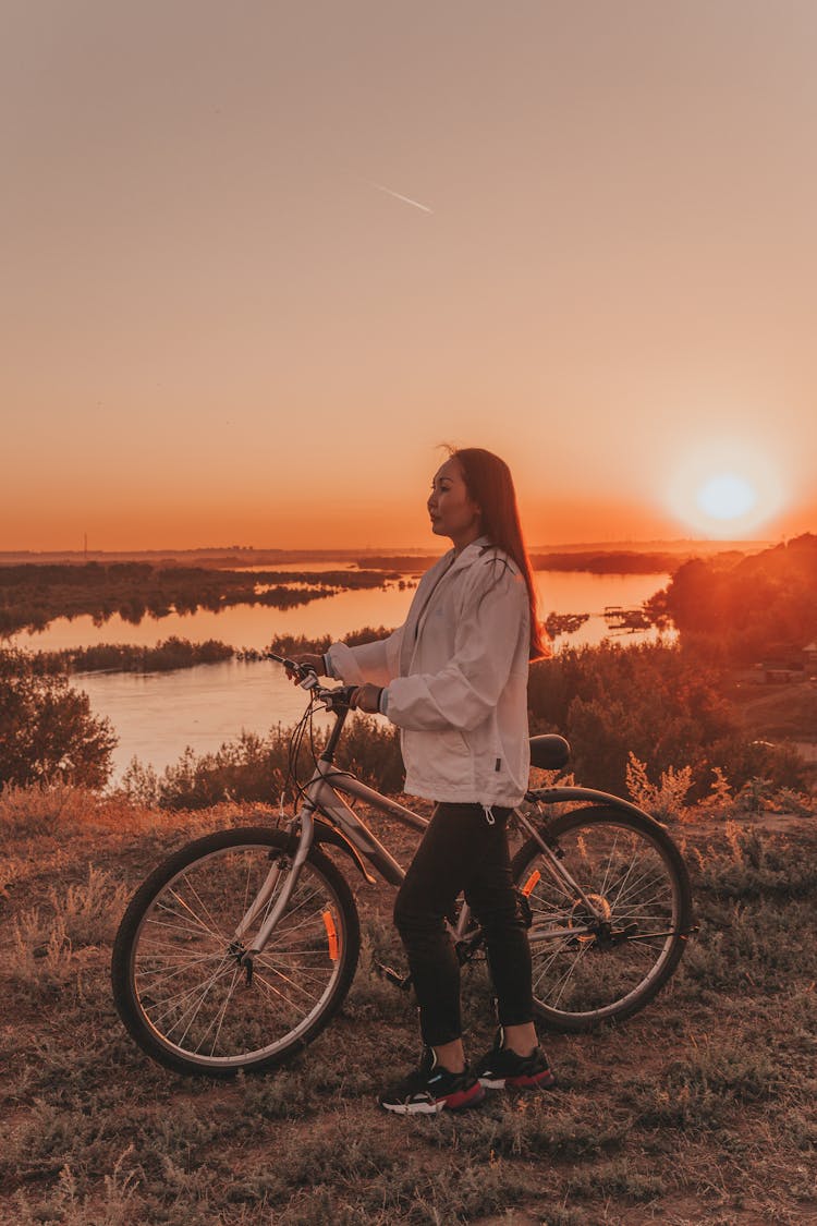 Asian Woman Standing Near Bicycle In Nature At Sunset