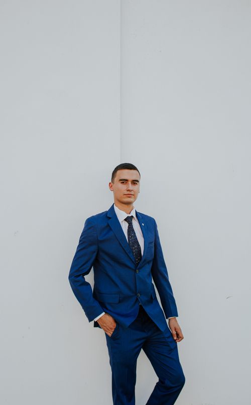 Free Young adult male confidently posing in a modern blue suit against a clean white wall. Stock Photo