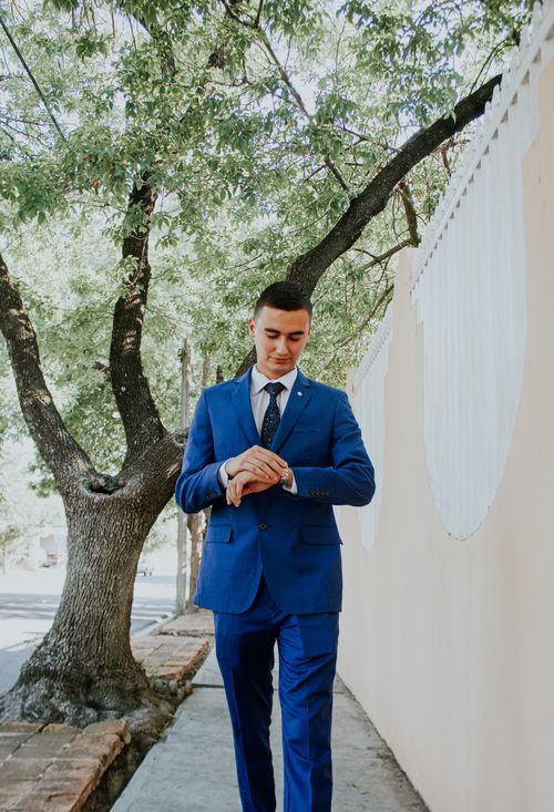 Free Man in blue suit walking on sidewalk, checking wristwatch. Stylish attire, outdoor setting. Stock Photo
