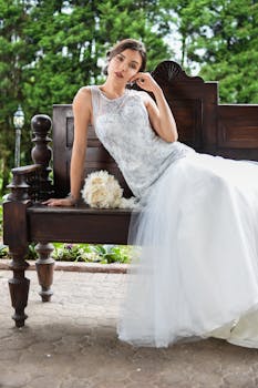 A bride in a stunning gown poses elegantly on a wooden bench outdoors, holding a bouquet.