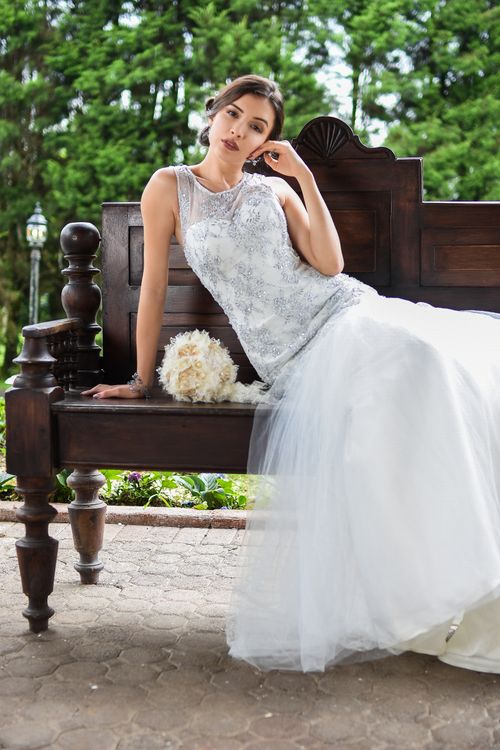 Free A bride in a stunning gown poses elegantly on a wooden bench outdoors, holding a bouquet. Stock Photo