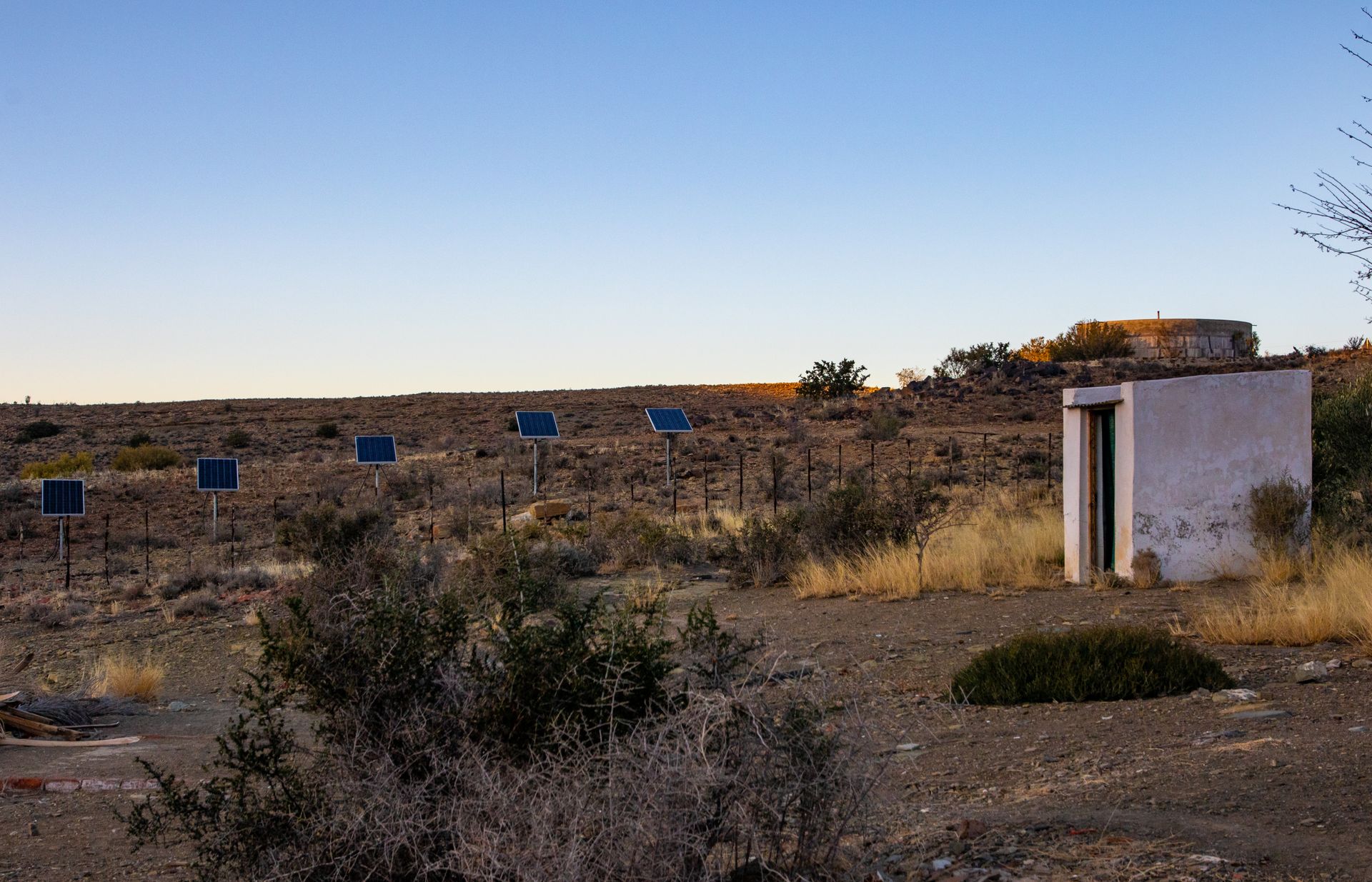 Solar panels in a desert landscape with a shed under a clear sunset sky.