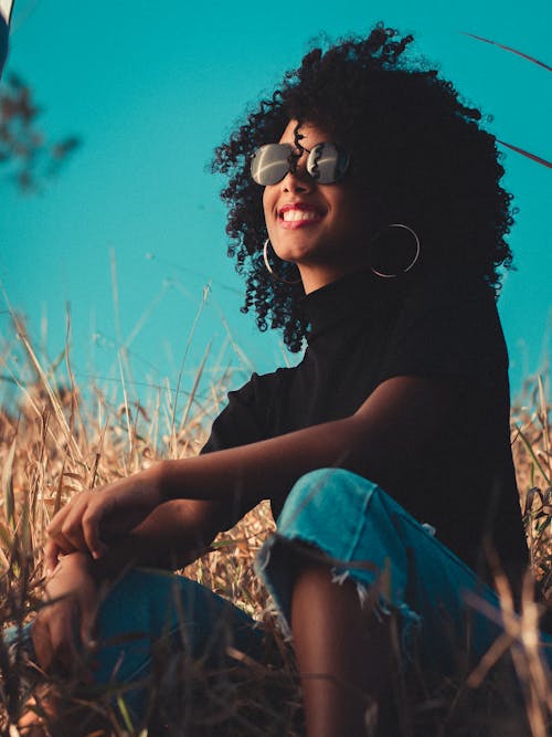 Charming young positive African American female in casual clothing and round sunglasses lounging in meadow with toothy smile under vibrant blue cloudless sky
