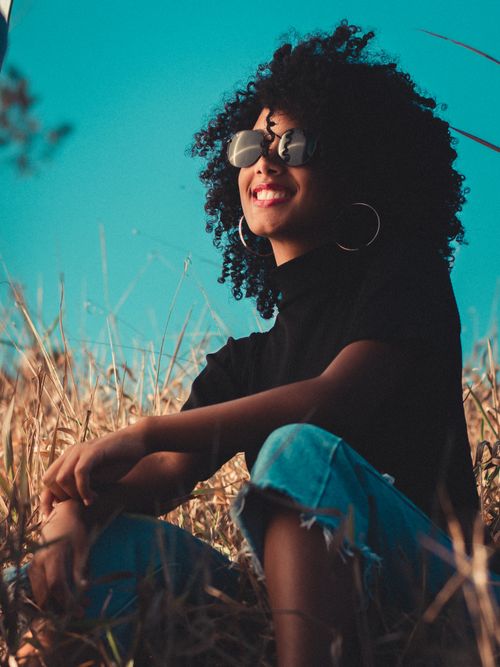 Free Charming young positive African American female in casual clothing and round sunglasses lounging in meadow with toothy smile under vibrant blue cloudless sky Stock Photo