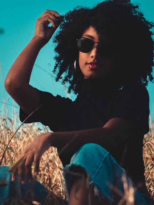 Dreamy young African American slender female in casual clothing and sunglasses touching curly locks while relaxing in meadow against blue sky