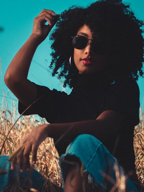 Free Dreamy young African American slender female in casual clothing and sunglasses touching curly locks while relaxing in meadow against blue sky Stock Photo