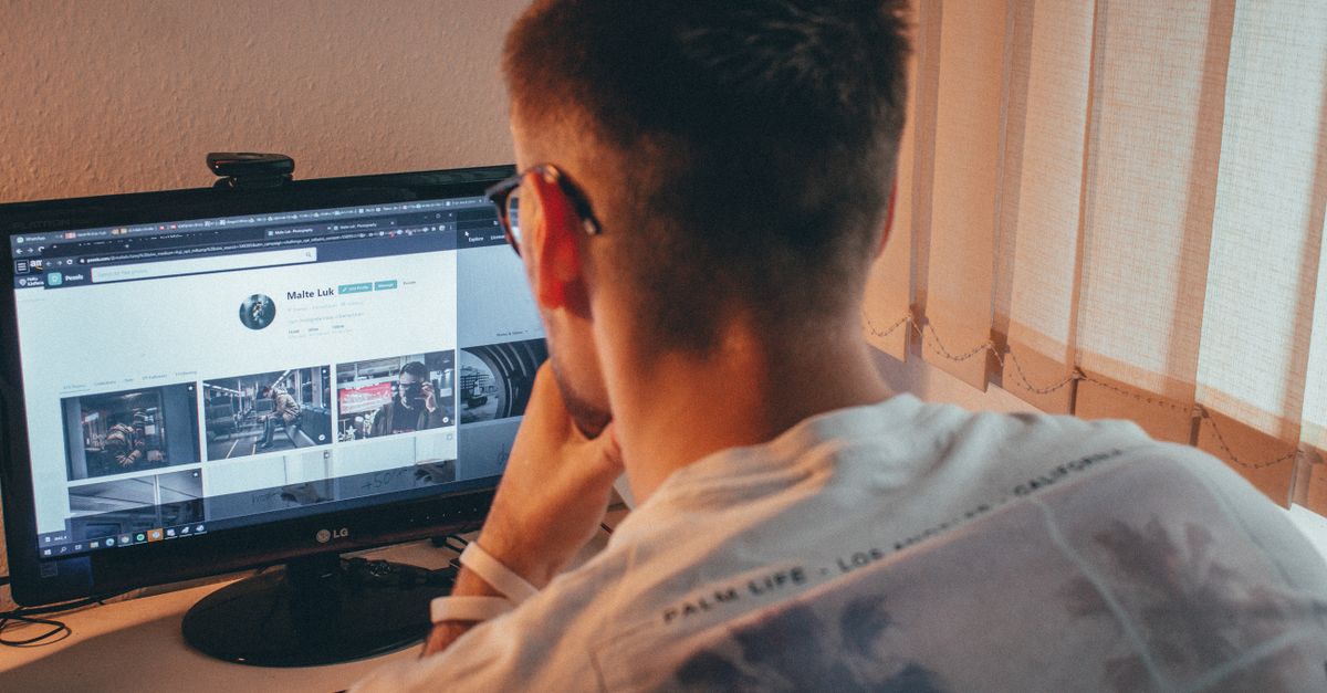 A young man wearing eyeglasses focused on browsing a website on his desktop computer indoors.