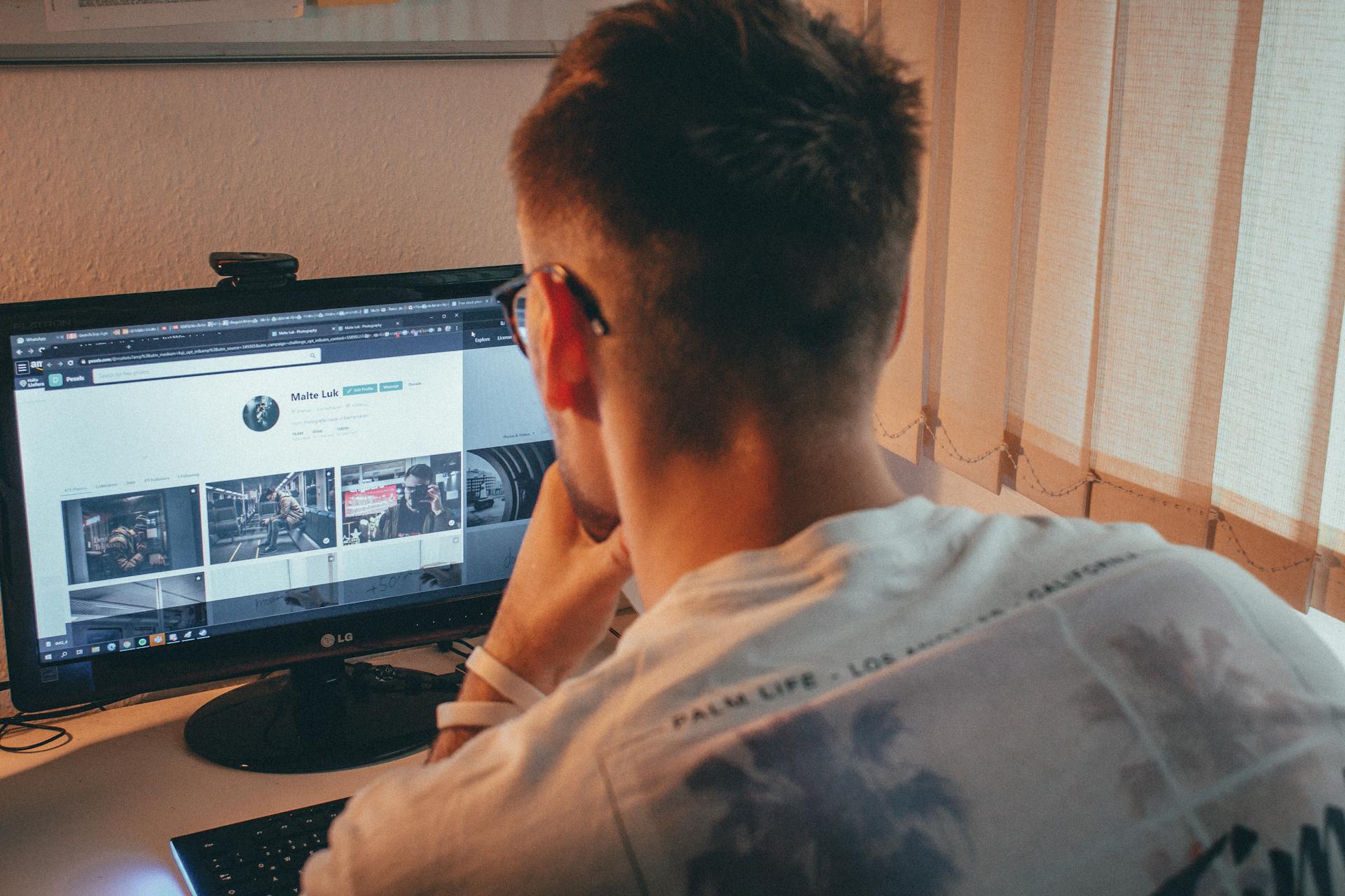 A young man wearing eyeglasses focused on browsing a website on his desktop computer indoors.