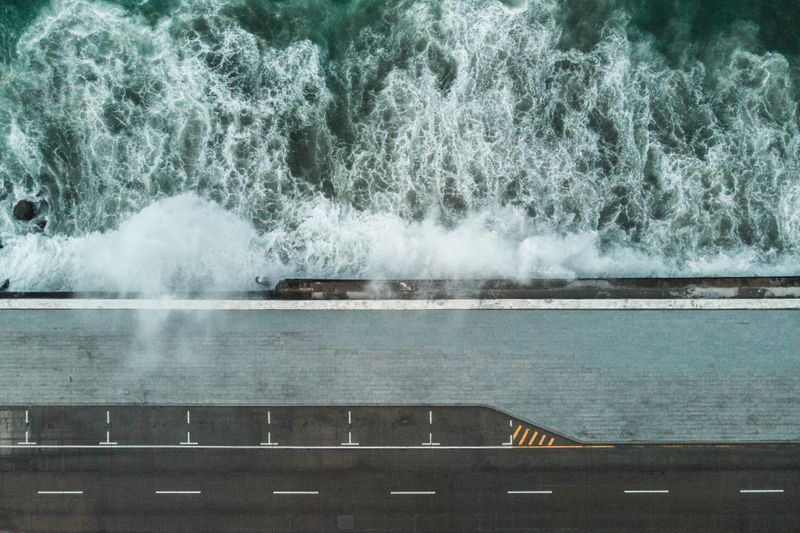 A dramatic aerial view capturing powerful ocean waves crashing against a coastal road.