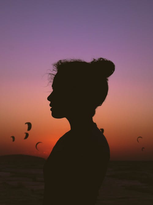 Free Side view silhouette of serene female standing against bright purple sunset sky with soaring paragliders Stock Photo