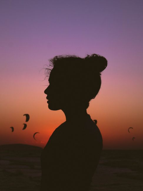 Side view silhouette of serene female standing against bright purple sunset sky with soaring paragliders