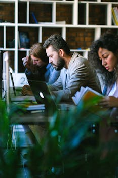 Thoughtful young colleagues in formal clothes sitting at desk with books and browsing netbooks while working in modern business center