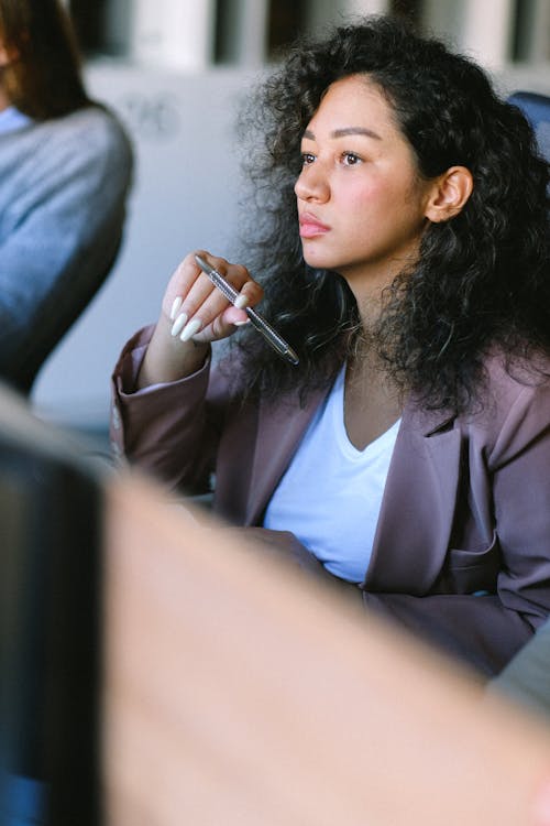 Free Young female employee wearing formal clothes sitting at table in office with pen and thoughtfully looking away Stock Photo