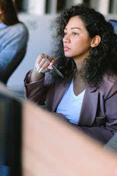 Young female employee wearing formal clothes sitting at table in office with pen and thoughtfully looking away