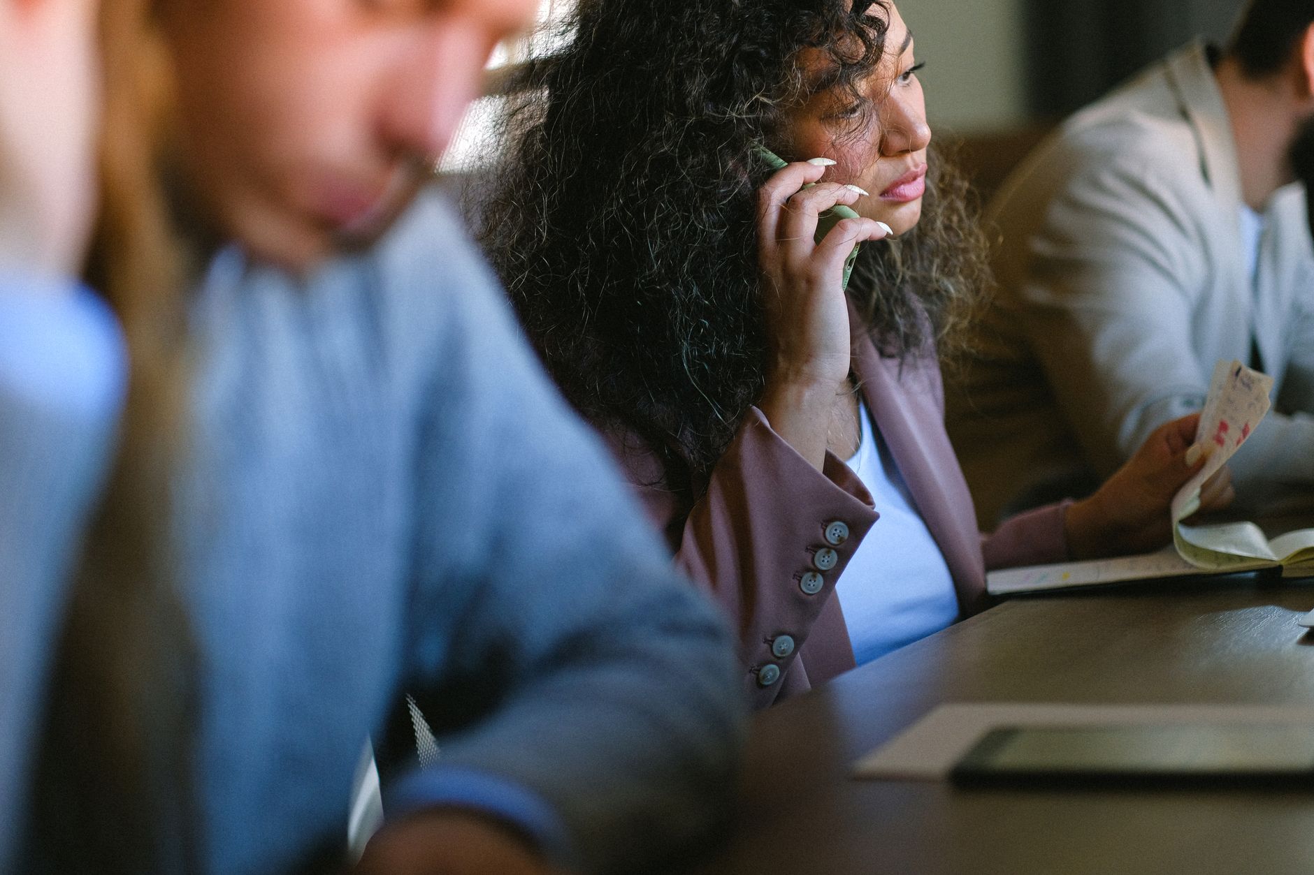 Concentrated female entrepreneur having phone call on cellphone while sitting at table with notebook with coworkers