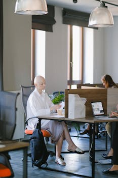 Full body of focused bald woman reading documents while sitting at table in open space with colleagues