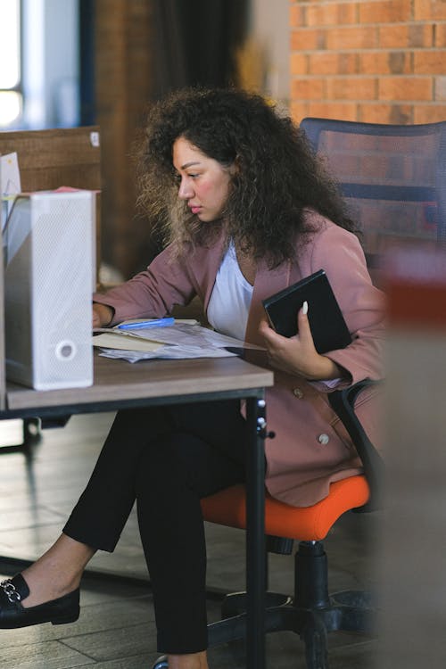Concentrated woman with curly hair in formal clothes working with documents at table in modern workplace