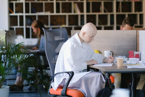 Side view of concentrated bald woman writing in paper document while working in office with colleagues