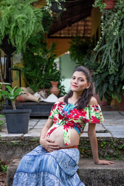 Free Positive young pregnant female in maxi dress and stylish top with bare shoulders and belly sitting on cottage stone steps and touching tummy while looking at camera Stock Photo