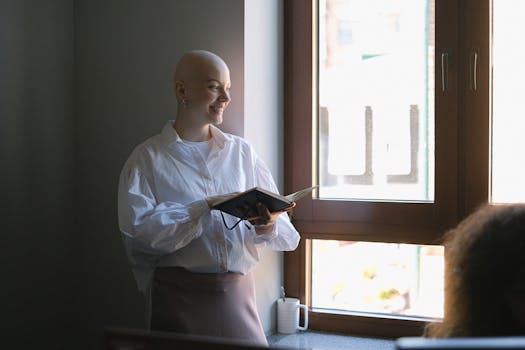 Positive bald female worker with notebook standing near window and explaining business project to coworker