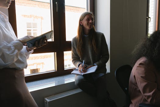 Positive male employee sitting on windowsill while discussing business issues with coworkers in office