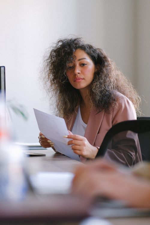 Free Pensive female with dark hair and paper sheet in hands sitting at table while working in conference room with blurred crop colleague Stock Photo