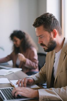 在现代办公室的模糊背景下，一位专注、留着胡须的男性经理正与同事一起工作，在上网本上打字