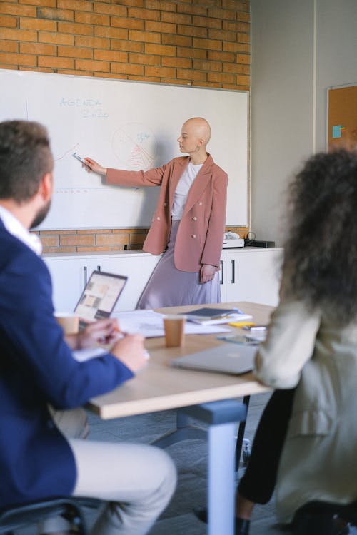 Busy bald female pointing with marker at board with diagrams while presenting new strategy to coworkers with laptop during meeting in office