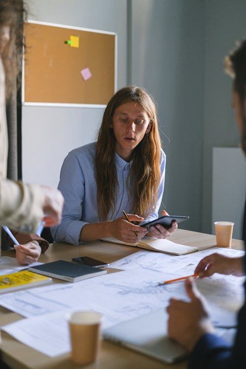 Free Concentrated male with long hair sitting at table with tablet and papers while working with coworkers during meeting in office Stock Photo