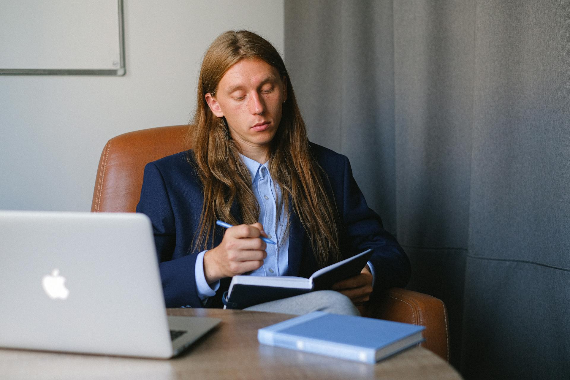 Pensive androgynous businessman in formal suit sitting at table with laptop and book and taking notes in notebook in workspace