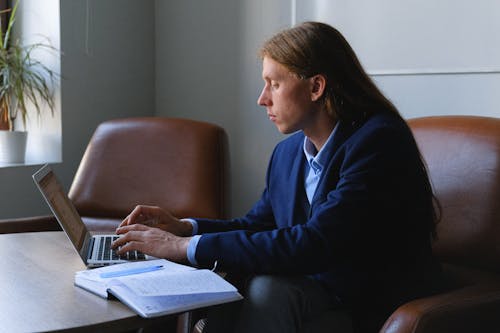 Side view of pensive androgynous entrepreneur with long hair in formal suit sitting at table with notebook and working on project on laptop in workspace in daytime