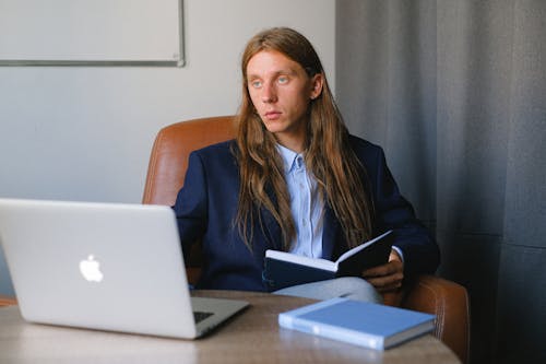 Free Serious entrepreneur in formal suit sitting at table with book and laptop while reading notebook and thoughtfully looking away in workspace in daytime Stock Photo