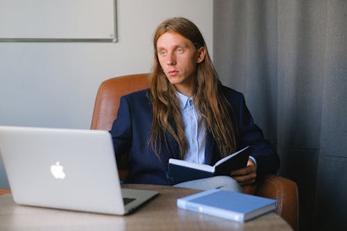 Serious entrepreneur in formal suit sitting at table with book and laptop while reading notebook and thoughtfully looking away in workspace in daytime