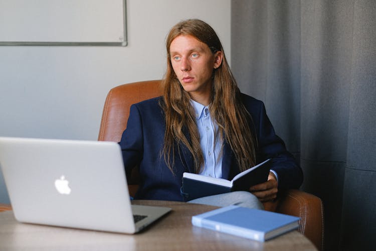 Androgynous Businessman Sitting At Table With Book And Laptop And Looking Away