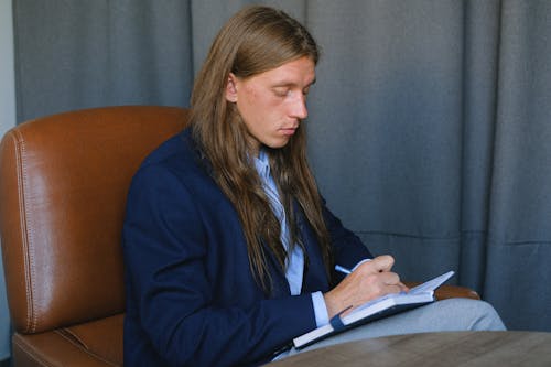 Pensive male with long hair writing notes in notebook
