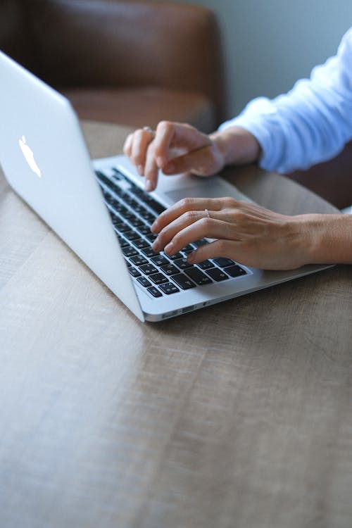 From above of crop faceless woman wearing pullover sitting at table and typing on modern laptop in daylight