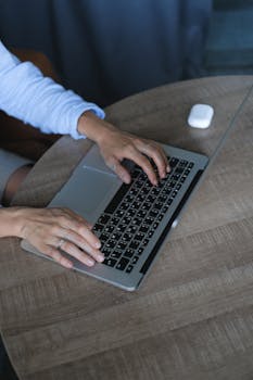 Close-up of a woman's hands typing on a laptop at a round wooden table indoors.
