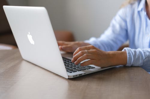 Crop unrecognizable lady wearing light blue shirt sitting at table with wireless computer and working remotely in daytime